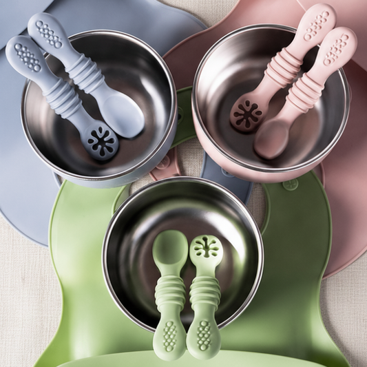 Three silicone baby feeding bibs in dusty blue, dusty pink, and sage green, each holding a stainless steel bowl and matching silicone spoon and teether, arranged on a white background.