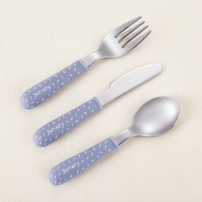 Three-piece baby cutlery set with silver stainless steel spoons and a fork and a lavender-blue plastic handles decorated with white polka dots and the word 'DAILY'.