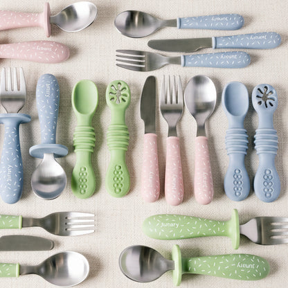 A set of pastel-colored baby cutlery with textured silicone handles, including pink, blue, and green utensils, arranged neatly on a white background.
