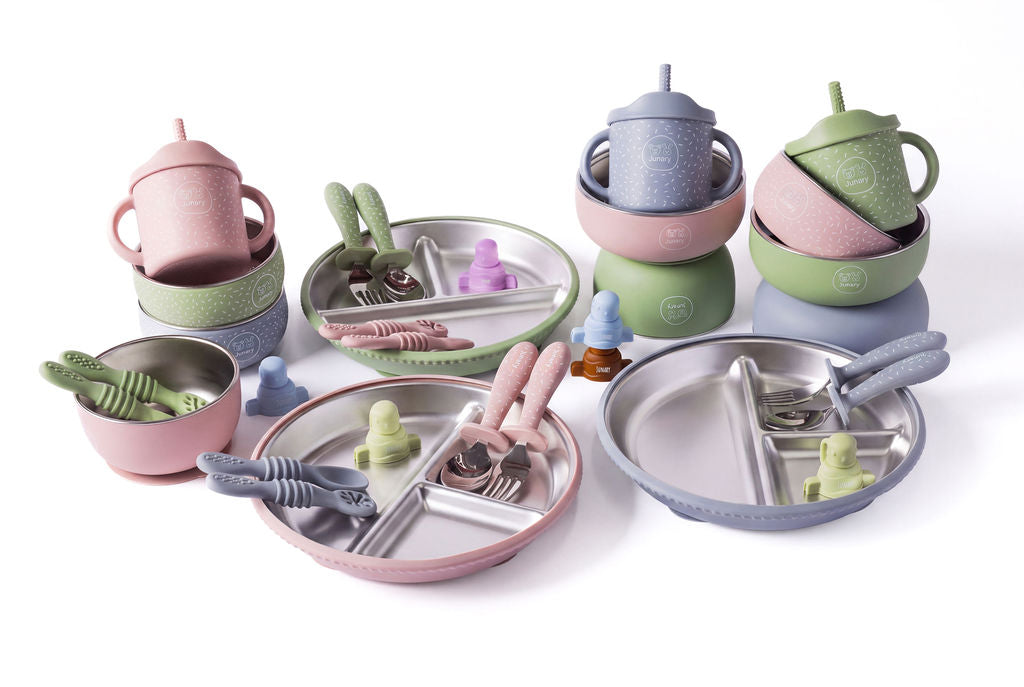 A coordinated children's dinnerware set arranged on a white studio background, including pastel pink, green, and blue plates, cups, bowls, and utensils with small frog-shaped accents.