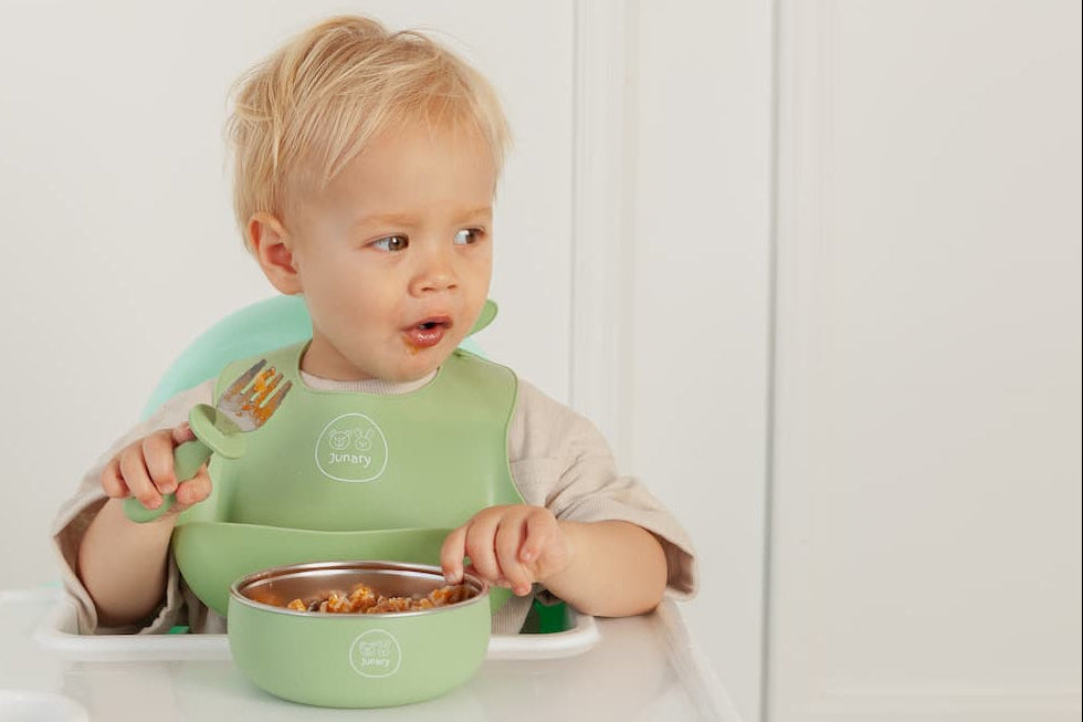 Child in a high chair with a green bib and bowl, eating.