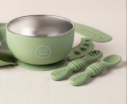 A baby feeding set in sage green silicone and stainless steel, including a suction cup bowl, a silicone bib, textured spoons, and a brush, arranged on a white background.