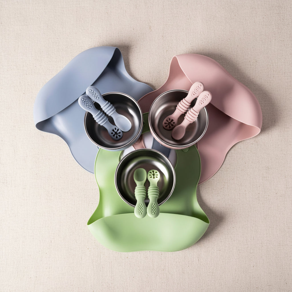 Three silicone baby feeding bibs in dusty blue, dusty pink, and light green, each holding a stainless steel bowl and matching silicone spoon set, arranged on a white background.