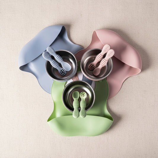 Three silicone baby feeding bibs in dusty blue, dusty pink, and light green, each holding a stainless steel bowl and matching silicone spoon set, arranged on a white background.