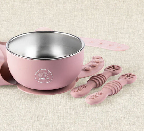 A baby feeding set in dusty pink silicone and stainless steel, including a bowl with a suction base, a silicone bib, and three textured silicone spoons, arranged on a white background.