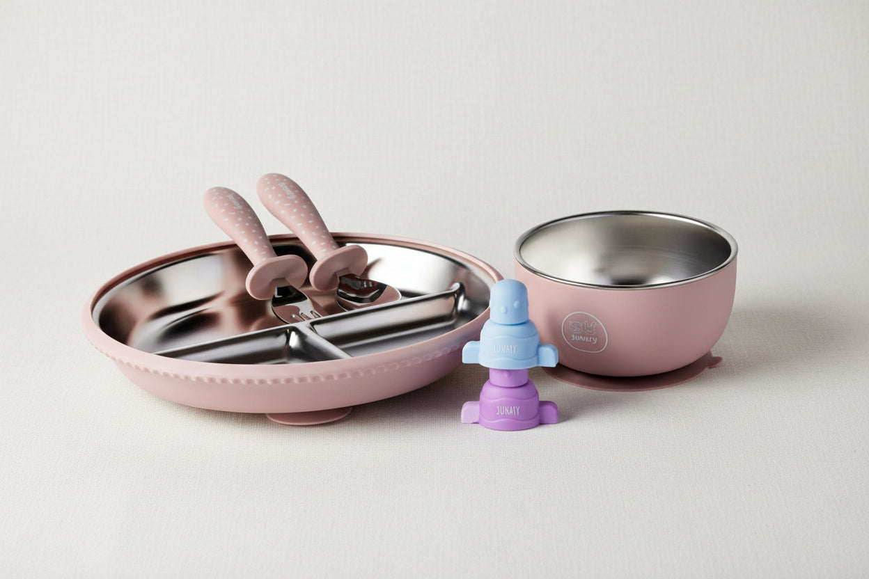 A baby feeding set featuring a pink plate with stainless steel utensils, a matching pink suction bowl with a stainless-steel interior, and a purple and blue pouch topper.