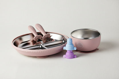 A baby feeding set featuring a pink plate with stainless steel utensils, a matching pink suction bowl with a stainless-steel interior, and a purple and blue pouch topper.